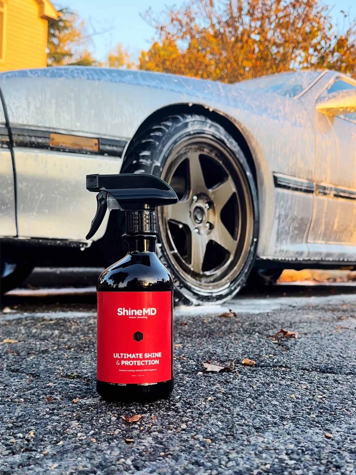 The ShineMD bottle staged on the driveway at golden hour, the car still rinsing off behind it — spray is the next step
